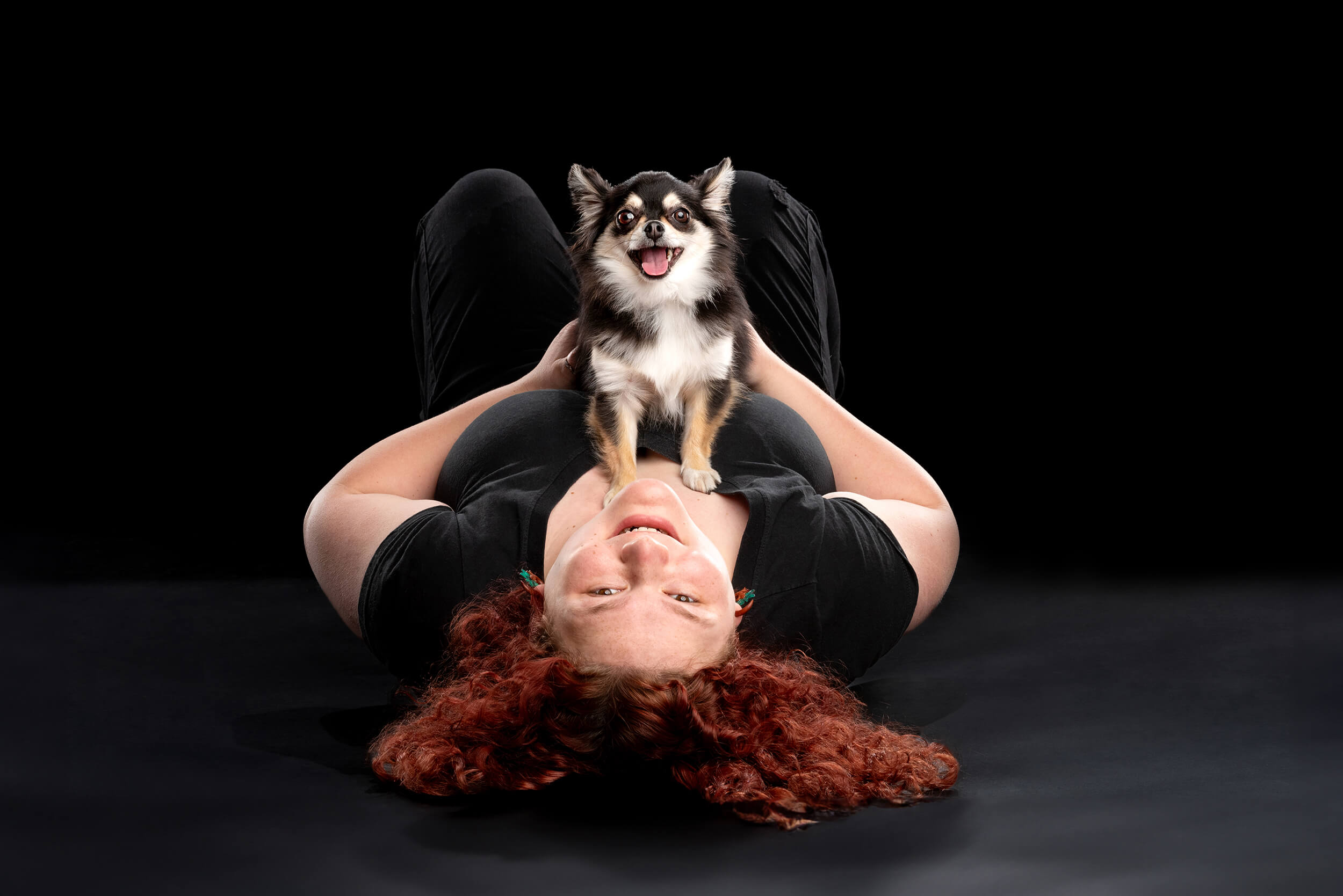 young adult with red curly hair laying with her small dog on top of her, in black clothing, black white and tan dog, by Chicago Pet Photographer Candice Cusic Photography